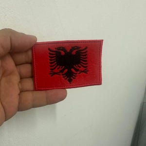 Op de afbeelding: Een rechthoekige rode patch met het zwarte dubbelkoppige adelaarsymbool van Albanië. De patch is waarschijnlijk gemaakt van stof en heeft een gestikte rand. De adelaar is gecentreerd op de patch.