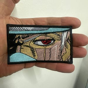 May include: Embroidered patch depicting an anime-style eye with a red iris and a tear. The rectangular patch has a black border, and the design incorporates shades of blue, tan, and white. The patch is held in a hand.