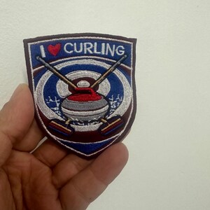Könnte beinhalten: Ein Aufnäher mit dem Text "I ❤️ CURLING" über einem Curling-Stein und gekreuzten Besen. Der Aufnäher hat eine dunkelburgunderfarbene Umrandung und einen blau-weißen Hintergrund.