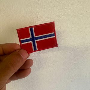 Può includere: Una toppa rettangolare con la bandiera norvegese. La bandiera è rossa con una croce nordica blu bordata di bianco. La toppa è probabilmente in tessuto ed è tenuta in mano.