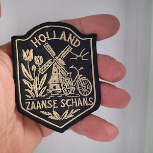 Può includere: Toppa ricamata nera e oro con la scritta "HOLLAND" sopra l'immagine di un mulino a vento, una bicicletta e le parole "ZAANSE SCHANS". La toppa include anche elementi floreali.
