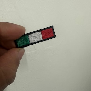 Può includere: Toppa ricamata rettangolare raffigurante la bandiera italiana. La toppa ha un bordo nero e mostra i colori della bandiera: verde, bianco e rosso. La toppa è piccola e sembra essere fatta di tessuto.