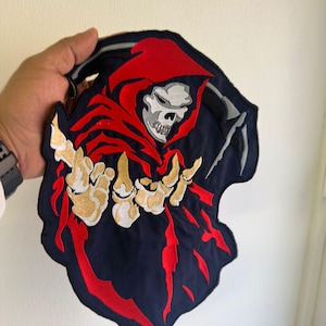 May include: Embroidered patch featuring a grim reaper design. The reaper is depicted with a white skull, a red hood, and gold-colored skeletal hands. The patch has a navy blue background with red accents and a black border.
