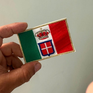 Può includere: Una toppa rettangolare ricamata che raffigura la bandiera storica d'Italia con strisce verticali verde, bianca e rossa. Al centro della striscia bianca si trova uno scudo rosso con croce bianca, sormontato da una corona e bordato d'oro.