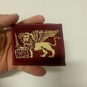 Può includere: Toppa bordeaux con un leone alato dorato che tiene una spada e un libro. Il leone è l'elemento centrale del design, con ali dettagliate e una posa regale. La toppa è rettangolare e ha una superficie strutturata.