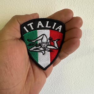 May include: A shield-shaped embroidered patch featuring the Italian flag colors of green, white, and red. The patch displays the Trinacria symbol in black and white, with the word ITALIA stitched in white capital letters along the top border.