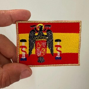 Può includere: Una toppa rettangolare ricamata raffigurante la storica bandiera spagnola con strisce orizzontali rosse e gialle. Al centro un'aquila nera con scudo, fiancheggiata da due colonne bianche con nastri rossi, il tutto bordato con filo dorato.