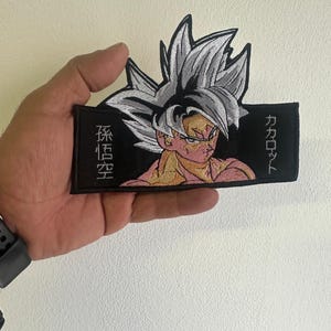 Può includere: Toppa ricamata raffigurante un personaggio con capelli argentati a spillo e un'espressione determinata. Il personaggio è ambientato su uno sfondo nero con testo giapponese. La toppa presenta un design dettagliato con colori vivaci.