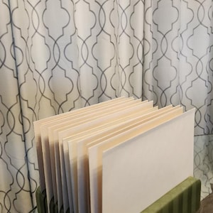 May include: A green, rectangular desk organizer with vertical stripes holds several cream-colored papers. The organizer sits on a reflective surface, with a patterned curtain in the background.