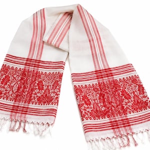 Pode incluir: Um cachecol retangular branco com padrões de riscas vermelhas e um intrincado bordado floral vermelho em ambas as extremidades. O tecido é rematado com delicadas franjas brancas e vermelhas nas extremidades inferiores, criando um design simétrico.