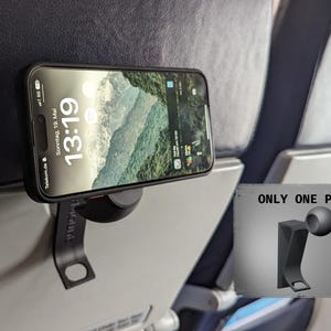 May include: A black smartphone is mounted on a black holder attached to an airplane seat. The phone displays a landscape image and the time 13:19. A small illustration on a gray sign reads "ONLY ONE PART."