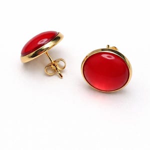 Puede incluir: Pendientes de botón con una piedra roja brillante de forma ovalada engastada en un bisel dorado. Los pendientes tienen un diseño clásico, con un cierre de poste y mariposa. El engaste dorado añade un toque de elegancia a la joya.