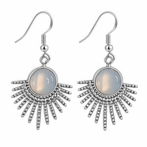 Pode incluir: Um par de brincos pendentes em tom prateado com uma pedra cabochão redonda branca translúcida no centro. A base de cada brinco tem um design de raios solares com hastes metálicas peroladas que se estendem a partir da pedra.