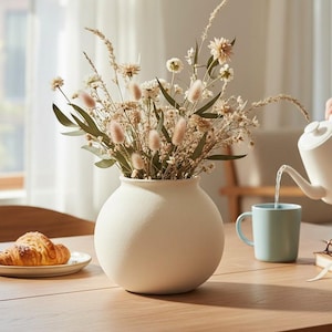 May include: A round, matte white ceramic vase filled with a bouquet of dried grasses and flowers, positioned on a wooden table next to a plate with a croissant, a light blue mug, a closed book, and a pair of round-rimmed glasses.