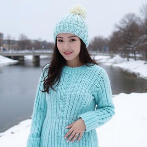 Könnte beinhalten: Hellblauer Strickpullover und passende Mütze mit weißem Bommel. Der Pullover hat ein Rippenmuster und lange Ärmel. Die Mütze ist im Zopfmuster-Stil gehalten. Die Frau steht draußen in einer verschneiten Umgebung.