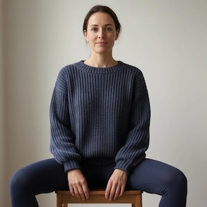 May include: A woman wearing a navy blue ribbed knit sweater and matching leggings. The sweater has a crew neck and long sleeves. She is sitting on a wooden stool in front of a plain white wall.