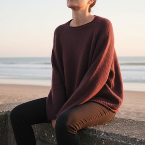 May include: A burgundy-colored sweater with a crew neck and long sleeves. The sweater is paired with black pants. The person is sitting on a concrete wall with the ocean and beach in the background. The sweater appears to be made of a soft, knitted material.