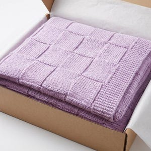 Puede incluir: Una manta de punto lavanda con un diseño tejido está doblada dentro de una caja de cartón. La manta está hecha de material suave y es de color morado claro. La caja está forrada con papel de seda blanco.