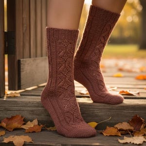 Puede incluir: Un par de calcetines de punto en un color rosa apagado, con un detallado patrón de punto de cable. Los calcetines se muestran sobre una superficie de madera con hojas de otoño caídas, lo que sugiere un tema acogedor y de temporada. Los calcetines están diseñados para la comodidad y el calor.
