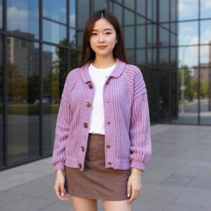 Puede incluir: Una mujer con una rebeca lila con cuello y botones marrones. La rebeca se combina con una camiseta blanca y una falda marrón. El fondo presenta un edificio moderno con ventanas de cristal.