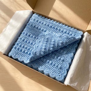 May include: A light blue, handmade crochet blanket with a scalloped edge, presented in a cardboard box with white tissue paper. The blanket features a textured stitch pattern. The overall theme is handmade baby items.