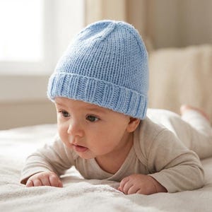 May include: A baby wearing a light blue knitted beanie hat and a beige onesie lies on a bed. The hat has a ribbed brim and a simple, classic design. The baby is looking off to the side.