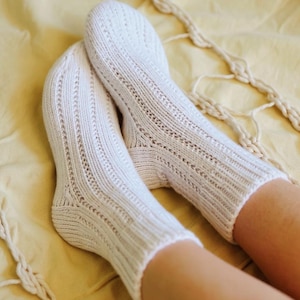 May include: A pair of cream-coloured knitted socks with a ribbed design and decorative openwork details. The socks are made of a soft, textured yarn and are shown on a yellow surface. These socks are a cosy and comfortable accessory.