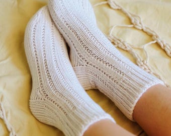 May include: A pair of off-white knitted socks with a ribbed design and decorative openwork details. The socks are made of a soft, textured yarn and are shown on a yellow surface. The socks are a cozy and comfortable accessory.