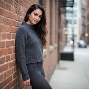 May include: A woman wearing a dark gray, cropped sweater with a ribbed hem and matching leggings. She is leaning against a red brick wall in an urban setting. The sweater has a high neckline.