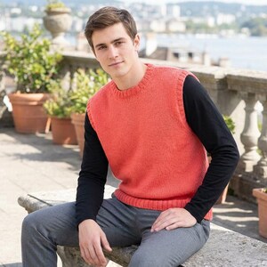 Könnte beinhalten: Eine korallenfarbene Strickweste über einem schwarzen Langarmshirt. Die Person trägt eine graue Hose und sitzt auf einer Steinbank. Der Hintergrund umfasst Topfpflanzen und einen Blick auf ein Gewässer.