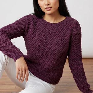May include: A plum-colored, textured knit sweater with a crew neck and long sleeves. The sweater has a woven pattern and is paired with white pants. The model is sitting on a wooden floor.
