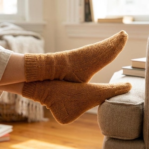 May include: A pair of knitted socks in a warm, golden-brown hue. The socks are resting on a neutral-toned sofa, showcasing their textured knit design. The socks appear to be comfortable and cozy, perfect for lounging.