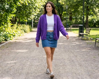 May include: A woman walks on a gravel path wearing a purple cardigan, white top, and denim skirt. The cardigan has a button-up front. She wears sneakers. The background includes trees, benches, and a green space.