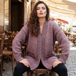 May include: A mauve-colored, open-front cardigan sweater with long sleeves and two front pockets. The sweater is made of a knitted material and is worn over a dark top and black pants. The background shows a cafe setting.
