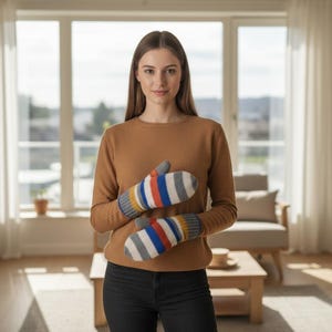 May include: A woman wearing a brown sweater and black jeans is holding a pair of mittens. The mittens are white with stripes of red, blue, yellow, and gray. The background is a well-lit living room.