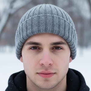 May include: A close-up portrait of a person wearing a gray ribbed knit beanie. The person is facing the camera with a neutral expression. The background is a blurred winter scene with snow and trees.
