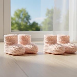 May include: Four pairs of light pink crochet baby booties are arranged on a light wood surface. The booties have a rolled cuff and a textured stitch pattern. The background includes a window with a view of trees and a sheer white curtain.