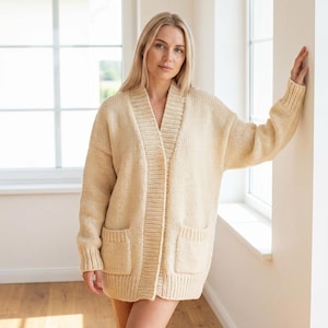 May include: A cream-colored, open-front cardigan sweater with two front pockets. The sweater has a wide collar and a relaxed fit. The model is wearing the sweater indoors, near a window.