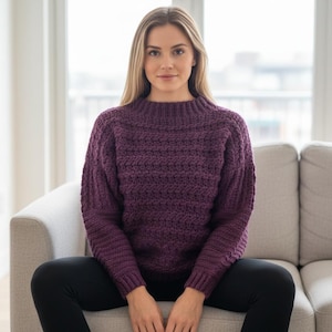 Pode incluir: Um suéter cor de ameixa com um padrão de malha texturizada. O suéter tem gola alta e canelada e mangas compridas. A pessoa está usando leggings pretas. O fundo inclui um sofá de tons neutros e uma janela.