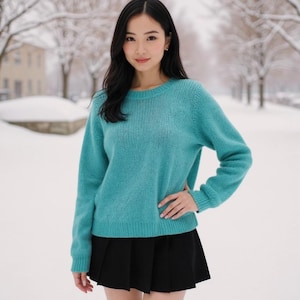 以下が含まれることがあります： 雪の降る屋外で、ターコイズ色のセーターと黒のプリーツスカートを着た人が立っています。セーターはクルーネックと長袖です。背景には雪に覆われた木々と建物があります。