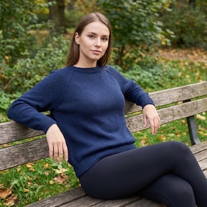 Könnte beinhalten: Eine Frau in einem marineblauen Pullover mit langen Ärmeln und schwarzen Leggings sitzt auf einer Holzbank im Park. Der Pullover hat einen Rundhalsausschnitt und Raglanärmel. Der Hintergrund zeigt Bäume und Blätter.