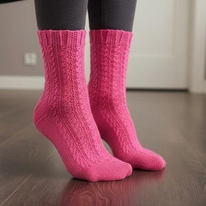 May include: A pair of bright pink knitted socks with a cable knit pattern. The socks are worn over dark gray leggings and are on a dark wood floor. The socks have a ribbed cuff and a textured design.