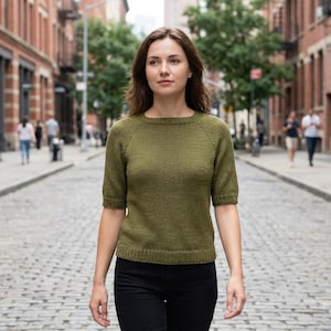 May include: An olive green knit top with short sleeves and a crew neck. The top is paired with black pants. The person is walking on a cobblestone street in a city setting.