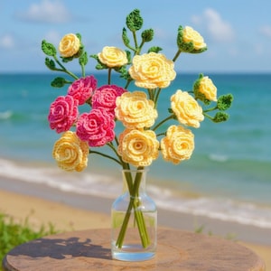 Puede incluir: Un ramo de rosas de ganchillo hechas a mano en un jarrón de cristal transparente. Las flores son amarillas y rosas, con hojas y tallos verdes. El jarrón está sobre una superficie de madera con una playa y un océano de fondo.