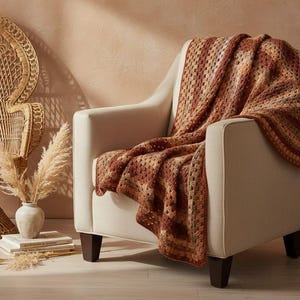 Puede incluir: Un sillón crema con una manta de ganchillo marrón y naranja sobre él. Una silla de pavo real tejida y un jarrón con hierba de la Pampa en el fondo crean una estética bohemia y acogedora.