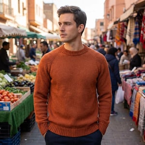 May include: A rust-colored crew neck sweater. The sweater has long sleeves and a ribbed neckline. The person is standing in a marketplace with produce and textiles. The sweater is made of a textured knit material.