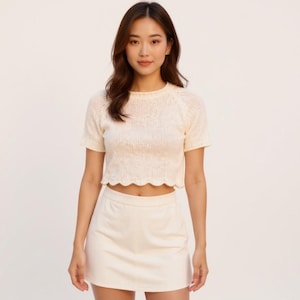 May include: A cream-coloured knitted crop top with a scalloped hem and short sleeves, worn with a matching mini skirt. The outfit is a light, neutral colour, suitable for a casual or semi-formal look.