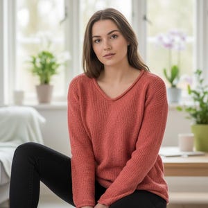 May include: A coral-colored, long-sleeved sweater with a V-neck. The sweater has a textured knit design. The person is wearing black pants. The background includes a window, plants, and a wooden table.