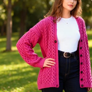 Könnte beinhalten: Ein leuchtend pinkfarbener Zopfstrick-Cardigan mit dunkelbraunen Knöpfen. Der Pullover hat ein strukturiertes Muster und lange Ärmel. Der Cardigan wird über einem weißen Oberteil und einer dunkelblauen Jeans mit einem schwarzen Gürtel getragen.