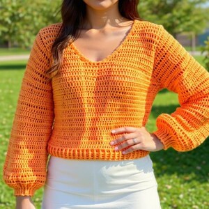 May include: Orange crochet sweater with a V-neck and long, puffy sleeves. The sweater has a textured, open weave design. The model is wearing a white skirt. The sweater is a bright, solid color.